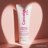 A white squeeze tube of Conceive Plus Fertility Lubricant with pink lettering, standing upright against a warm pink background with soft heart-shaped shadows.