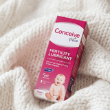 A box of Conceive Plus Fertility Lubricant resting on a soft, white chunky knit blanket. The packaging is predominantly pink and white with text indicating it supports sperm quality and is suitable for pregnant women.