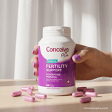 A hand with purple nail polish holds a white bottle of Conceive Plus Women's Fertility Support dietary supplement, with pink capsules scattered on a light wooden surface in front of it.